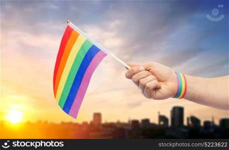 lgbt, same-sex relationships and homosexual concept - close up of male hand wearing gay pride awareness wristband holding rainbow flag over city sunset background. hand with gay pride rainbow flag and wristband. hand with gay pride rainbow flag and wristband