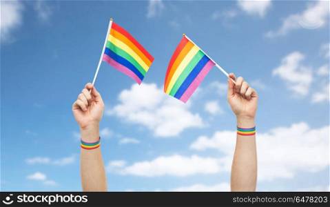lgbt, same-sex relationships and homosexual concept - close up of male hand wearing gay pride awareness wristbands holding rainbow flags over blue sky and clouds background. hand with gay pride rainbow flags and wristbands. hand with gay pride rainbow flags and wristbands