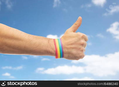 lgbt, same-sex relationships and homosexual concept - close up of male hand wearing gay pride awareness wristband showing thumbs up over blue sky and clouds background. hand with gay pride rainbow wristband shows thumb. hand with gay pride rainbow wristband shows thumb