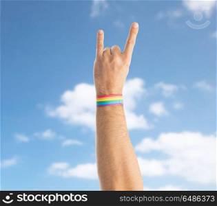lgbt, same-sex relationships and homosexual concept - close up of male hand wearing gay pride awareness wristband showing rock or hand-horns sign over blue sky and clouds background. hand with gay pride rainbow wristband shows rock. hand with gay pride rainbow wristband shows rock