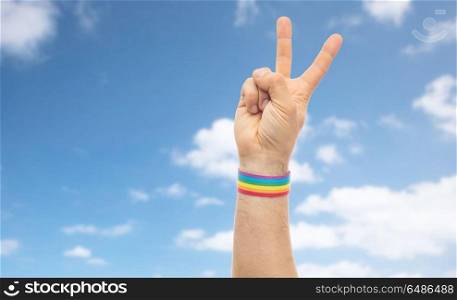 lgbt, same-sex relationships and homosexual concept - close up of male hand wearing gay pride awareness wristband showing peace sign over blue sky and clouds background. hand with gay pride rainbow wristband make peace. hand with gay pride rainbow wristband make peace