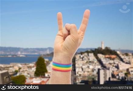 lgbt, same-sex relationships and homosexual concept - close up of male hand wearing gay pride awareness wristband showing rock or hand-horns sign over san francisco city background. hand with gay pride rainbow wristband shows rock. hand with gay pride rainbow wristband shows rock