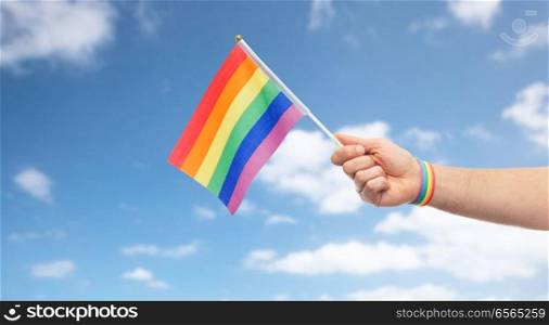 lgbt, same-sex relationships and homosexual concept - close up of male hand with gay pride awareness wristband holding rainbow flag over blue sky and clouds background. hand with gay pride rainbow flag and wristband