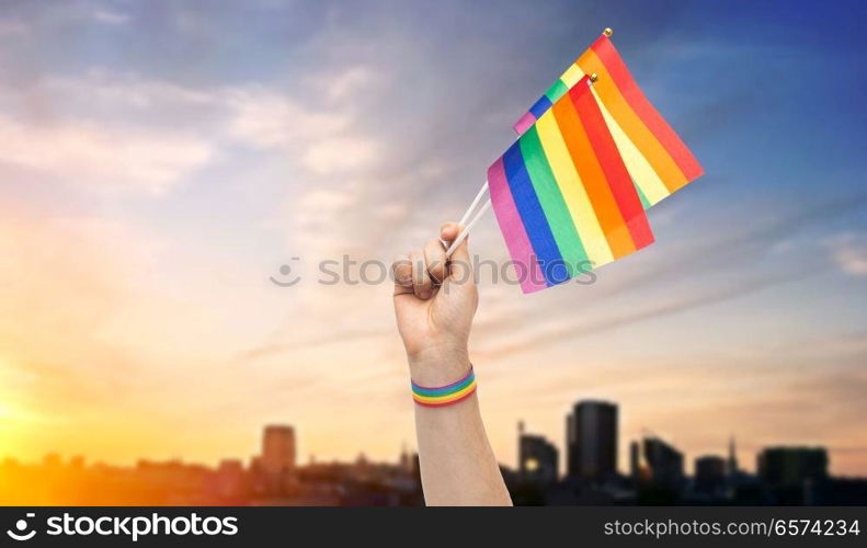 lgbt, same-sex relationships and homosexual concept - close up of male hand with gay pride awareness wristband holding rainbow flags over sunset in tallinn city background. hand with gay pride rainbow flags and wristband