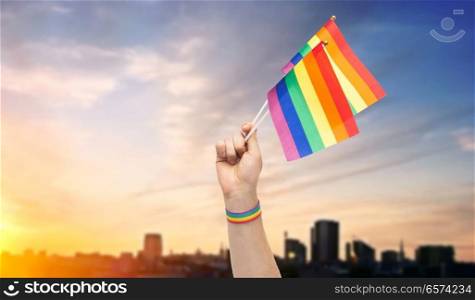 lgbt, same-sex relationships and homosexual concept - close up of male hand with gay pride awareness wristband holding rainbow flags over sunset in tallinn city background. hand with gay pride rainbow flags and wristband