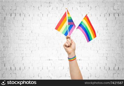 lgbt, same-sex relationships and homosexual concept - close up of male hand wearing gay pride awareness wristband holding rainbow flags over brick wall background. hand with gay pride rainbow flags and wristband
