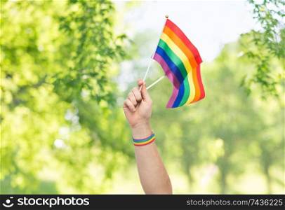 lgbt, same-sex relationships and homosexual concept - close up of male hand wearing gay pride awareness wristband holding rainbow flags over green natural background. hand with gay pride rainbow flags and wristband