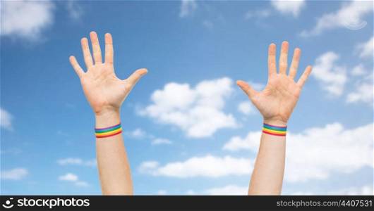 lgbt, same-sex relationships and homosexual concept - close up of male hands wearing gay pride awareness wristbands over blue sky and clouds background. hands with gay pride rainbow wristbands. hands with gay pride rainbow wristbands