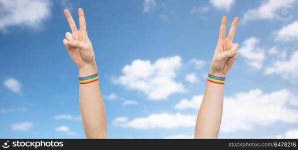 lgbt, same-sex relationships and homosexual concept - close up of male hands wearing gay pride awareness wristbands showing peace sign over blue sky and clouds background. hands with gay pride rainbow wristbands make peace. hands with gay pride rainbow wristbands make peace