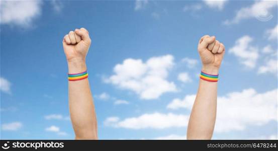 lgbt, same-sex relationships and homosexual concept - close up of male hands wearing gay pride awareness wristbands showing fist over blue sky and clouds background. hands with gay pride rainbow wristbands shows fist. hands with gay pride rainbow wristbands shows fist