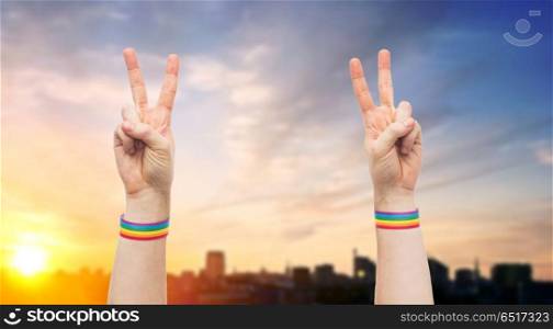 lgbt, same-sex relationships and homosexual concept - close up of male hands wearing gay pride awareness wristbands showing peace sign over city sunset background. hands with gay pride rainbow wristbands make peace. hands with gay pride rainbow wristbands make peace