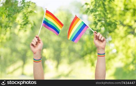 lgbt, same-sex relationships and homosexual concept - close up of male hands wearing gay pride awareness wristbands holding rainbow flags over green natural background. hands with gay pride rainbow flags and wristbands