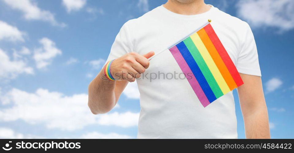 lgbt, same-sex relationships and homosexual concept - close up of man wearing gay pride awareness wristband holding rainbow flag over blue sky and clouds background. man with gay pride rainbow flag and wristband. man with gay pride rainbow flag and wristband