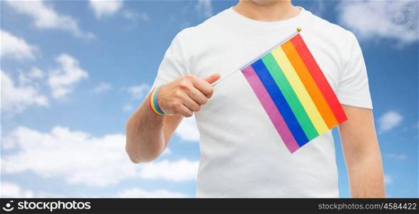 lgbt, same-sex relationships and homosexual concept - close up of man wearing gay pride awareness wristband holding rainbow flag over blue sky and clouds background. man with gay pride rainbow flag and wristband. man with gay pride rainbow flag and wristband