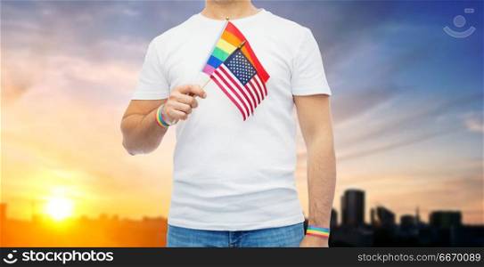 lgbt, same-sex relationships and homosexual concept - close up of man wearing gay pride rainbow awareness wristbands and holding american flag over city sunset background. gay man with american flag and rainbow wristbands. gay man with american flag and rainbow wristbands