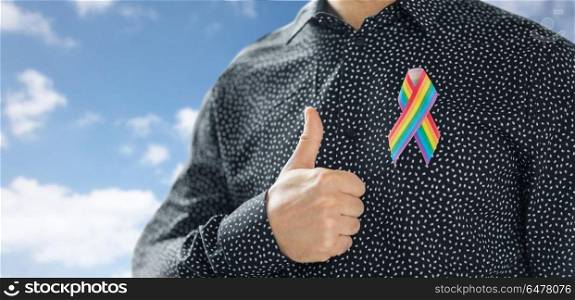lgbt, same-sex relationships and homosexual concept - close up of man with gay pride rainbow awareness ribbon on his chest showing thumbs up over blue sky and clouds background. man with gay pride rainbow awareness ribbon. man with gay pride rainbow awareness ribbon