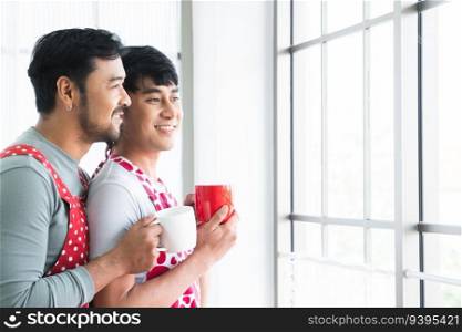 Lgbtq gay couple handsome men hugging from back and holding cup of coffee, smiling looking out of window in morning in kitchen at home while have breakfast. Valentines day concept