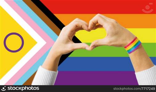 lgbtq, trans and intersex rights concept - close up of couple wearing rainbow ribbon wristbands and showing hand heart over progress pride flag on background. couple showing hand heart over progress pride flag