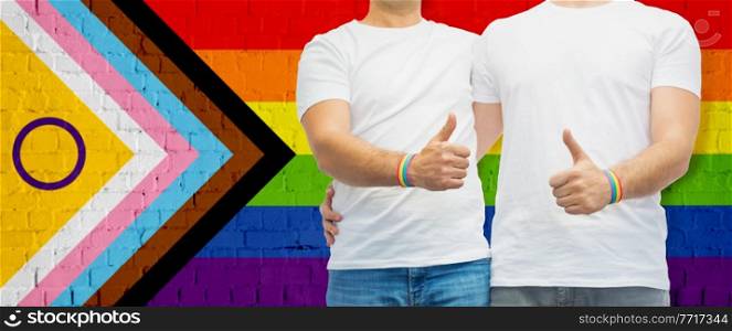 lgbtq, trans and intersex rights concept - close up of male gay couple hugging and showing thumbs up over rainbow progress pride flag on background. close up of male gay couple over pride flag