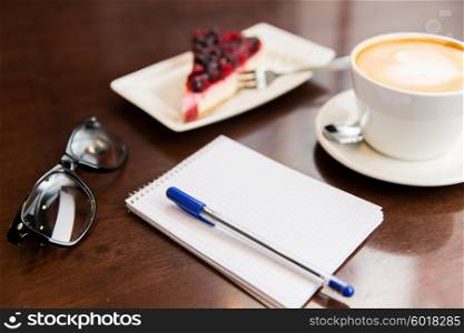 lifestyle and object concept - close up of notebook with pen, eyeglasses, coffee cup and berry cake on table