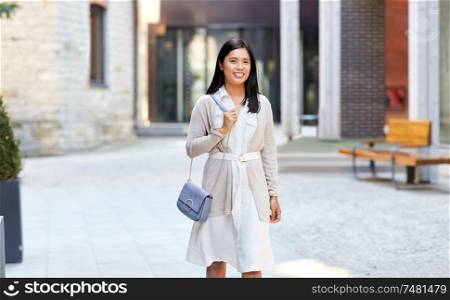 lifestyle and people concept - happy smiling young asian woman with handbag walking along city street. happy smiling young asian woman on city street