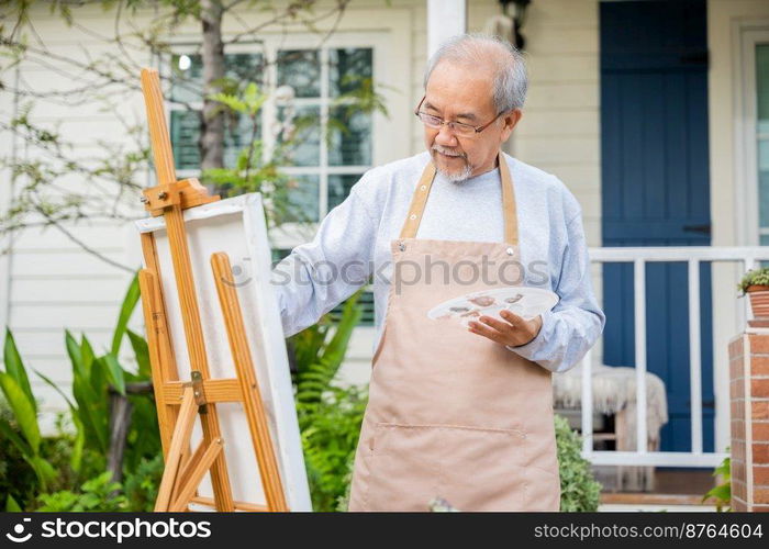 Lifestyle Asian senior old man painting picture artwork using brush and oil color on canvas, elderly people smile paint at his easel outside green nature background, Happy retirement artist activity