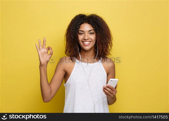Lifestyle Concept - Portrait of beautiful African American woman joyful listening to music on mobile phone and show ok sign with fingers. Yellow pastel studio background. Copy Space. Lifestyle Concept - Portrait of beautiful African American woman joyful listening to music on mobile phone and show ok sign with fingers. Yellow pastel studio background. Copy Space.