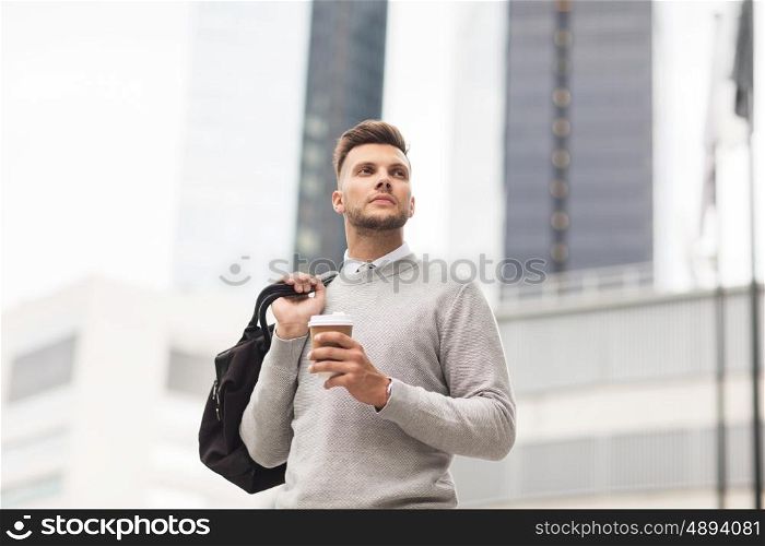 lifestyle, drinks and people concept - young man with bag drinking coffee from paper cup in city