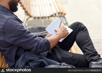 lifestyle, freelance, inspiration and people concept - close up of man with bag writing to notebook or diary sitting on city street bench