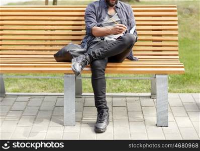 lifestyle, freelance, inspiration and people concept - close up of man with bag writing to notebook or diary sitting on city street bench
