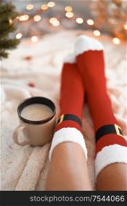 Lifestyle home photo of feet in Santa&rsquo;s socks near the Christmas tree. Woman sitting at the blanket, drinks hot beverage and relaxes warming up their feet in woollen socks. Winter and Christmas holidays concept