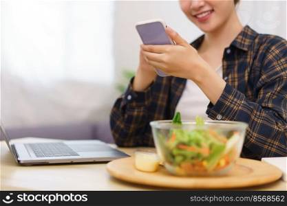 Lifestyle in living room concept, Young Asian woman using smartphone and eating vegetable salad.