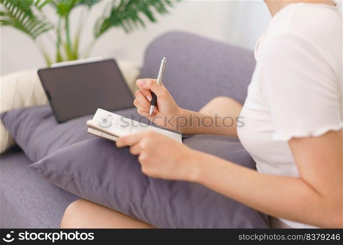 Lifestyle in living room concept, Young Asian woman writing data on notebook while sitting on couch.