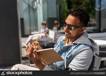 lifestyle, inspiration and people concept - indian man writing to notebook or diary at street cafe. man with notebook and coffee at street cafe