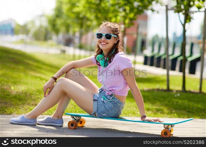 lifestyle, leisure and people concept - smiling young woman or teenage girl in sunglasses with headphones sitting on longboard in park. happy teenage girl with headphones and longboard