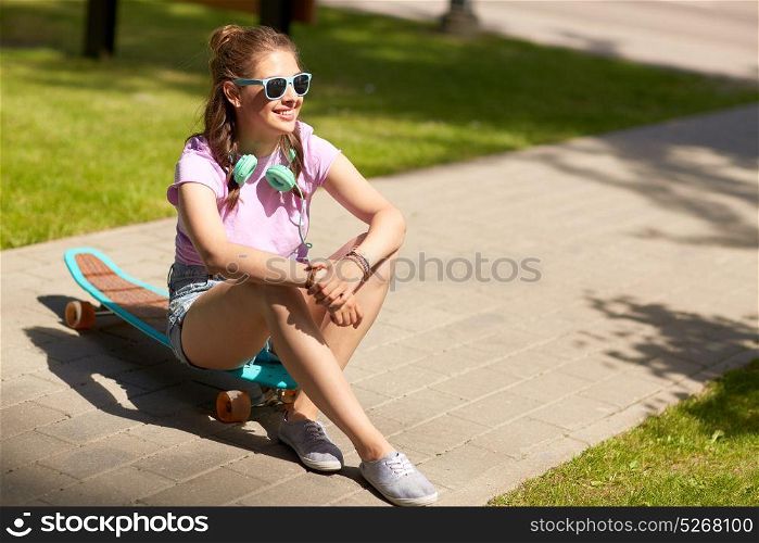 lifestyle, leisure and people concept - smiling young woman or teenage girl in sunglasses with headphones sitting on longboard in park. happy teenage girl with headphones and longboard