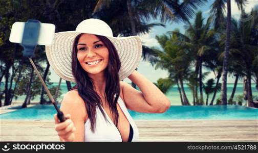 lifestyle, leisure, summer, technology and people concept - smiling young woman in sun hat taking picture with smartphone on selfie stick over tropical beach with palms and swimming pool background