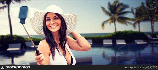 lifestyle, leisure, summer, technology and people concept - smiling young woman in sun hat taking picture with smartphone on selfie stick over resort beach with palms and swimming pool background