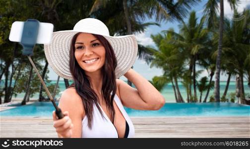 lifestyle, leisure, summer, technology and people concept - smiling young woman in sun hat taking picture with smartphone on selfie stick over tropical beach with palms and swimming pool background