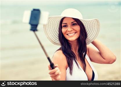 lifestyle, leisure, summer, technology and people concept - smiling young woman or teenage girl in sun hat taking picture with smartphone on selfie stick at beach