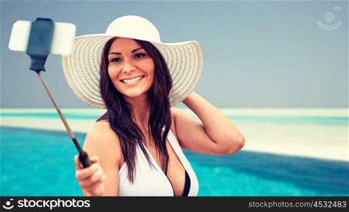 lifestyle, leisure, summer, technology and people concept - smiling young woman or teenage girl in sun hat taking picture with smartphone on selfie stick over beach and swimming pool background