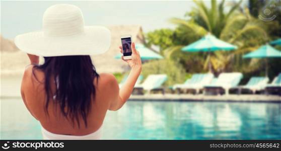 lifestyle, leisure, summer, technology and people concept - smiling young woman or teenage girl in sun hat taking selfie with smartphone over beach and swimming pool background