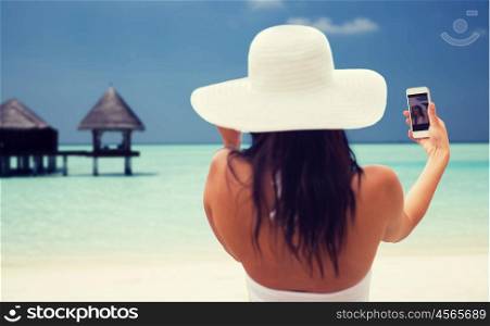 lifestyle, leisure, summer, technology and people concept - smiling young woman or teenage girl in sun hat taking selfie with smartphone over bungalow on beach background
