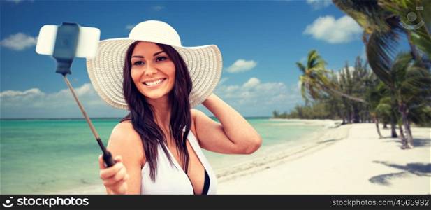 lifestyle, leisure, summer, technology and people concept - smiling young woman or teenage girl in sun hat taking picture with smartphone on selfie stick over tropical beach with palms background