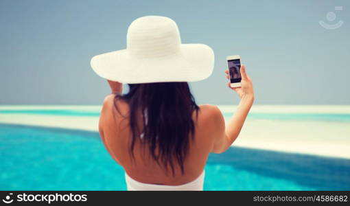 lifestyle, leisure, summer, technology and people concept - smiling young woman or teenage girl in sun hat taking selfie with smartphone over beach and swimming pool background