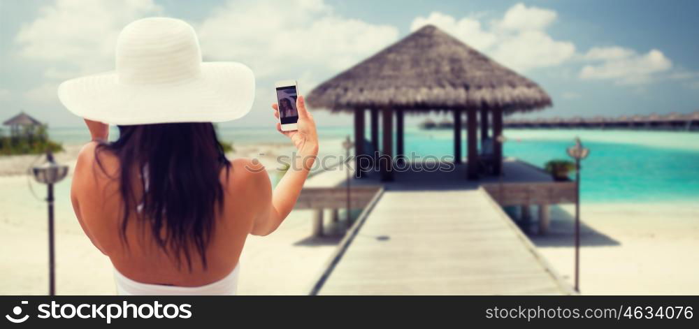 lifestyle, leisure, summer, technology and people concept - smiling young woman or teenage girl in sun hat taking selfie with smartphone over bungalow on beach background