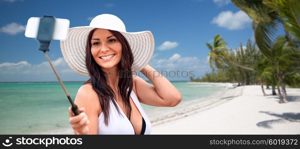 lifestyle, leisure, summer, technology and people concept - smiling young woman or teenage girl in sun hat taking picture with smartphone on selfie stick over tropical beach with palms background