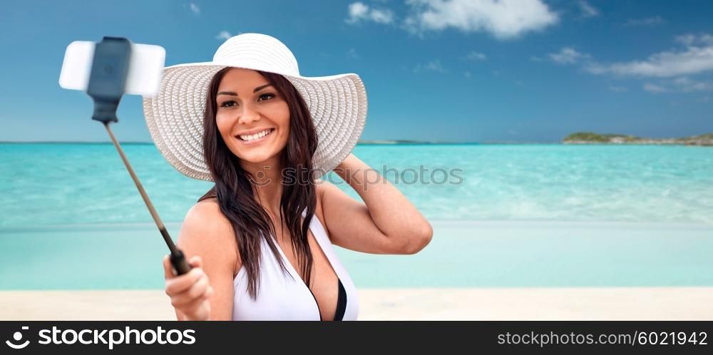 lifestyle, leisure, summer, technology and people concept - smiling young woman or teenage girl in sun hat taking picture with smartphone on selfie stick over tropical beach background