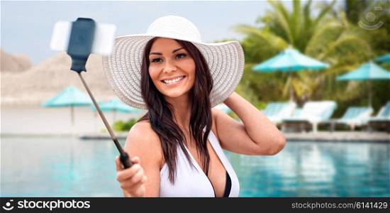 lifestyle, leisure, summer, technology and people concept - smiling young woman or teenage girl in sun hat taking picture with smartphone on selfie stick over beach and swimming pool background