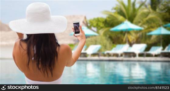 lifestyle, leisure, summer, technology and people concept - smiling young woman or teenage girl in sun hat taking selfie with smartphone over beach and swimming pool background
